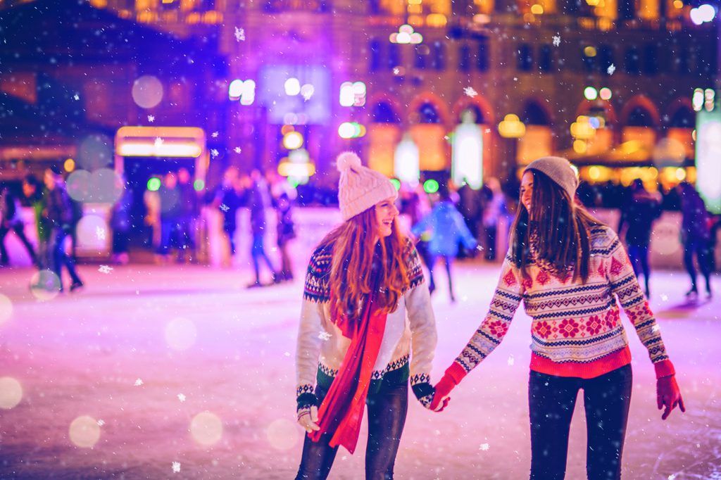 Friends having so much fun while ice skating at night. Wearing warm clothing. City is decorated with christmas lights.