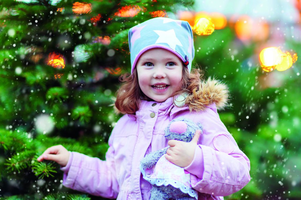 Cute little smiling kid girl with christmas tree. Happy child in winter clothes and toy choosing xmas tree on Christmas market with lights on background. Family, tradition, celebration concept