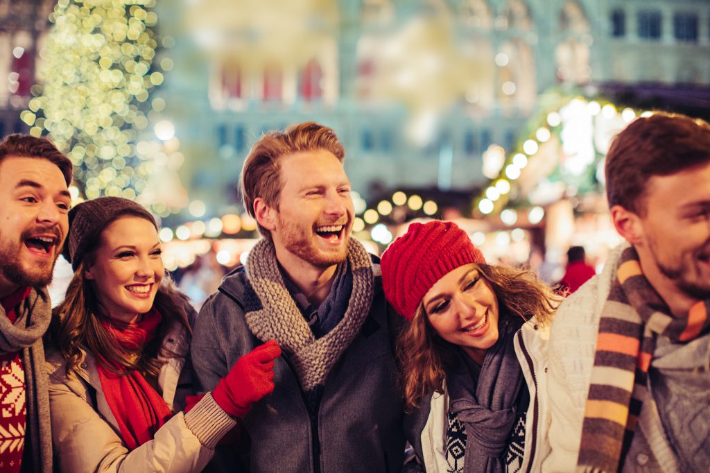 Young group of friends having fun outdoors at winter. Wearing warm clothes, hats and scarfs.Visiting christmas market in Vienna, Austria. Everything is decorated with festive string lights. Evening or night with beautiful yellow lights lightning the scenes.