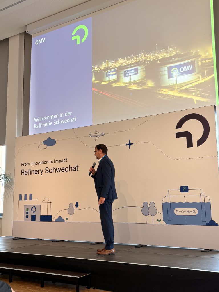 Otmar Schneider spricht auf der Bühne vor einer Projektion mit dem Schriftzug Willkommen in der Raffinerie Schwechat